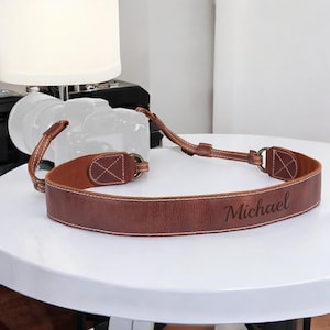 May include: A brown leather camera strap with white stitching and metal hardware. The name "Michael" is engraved on the strap. The strap is designed to be worn around the neck.
