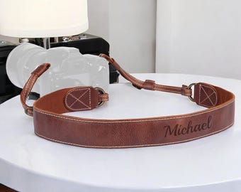 Może przedstawiać: Brązowy skórzany pasek do aparatu z białymi przeszyciami i metalowymi elementami. Na pasku wygrawerowano imię "Michael". Pasek jest przeznaczony do noszenia na szyi.