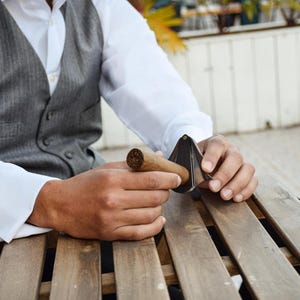 Puede incluir: Un cortapuros met&aacute;lico en forma de V con acabado plateado, colocado para cortar el extremo de un cigarro de tabaco marr&oacute;n. La herramienta presenta un dise&ntilde;o articulado con un punto de pivote central para preparar el cigarro.