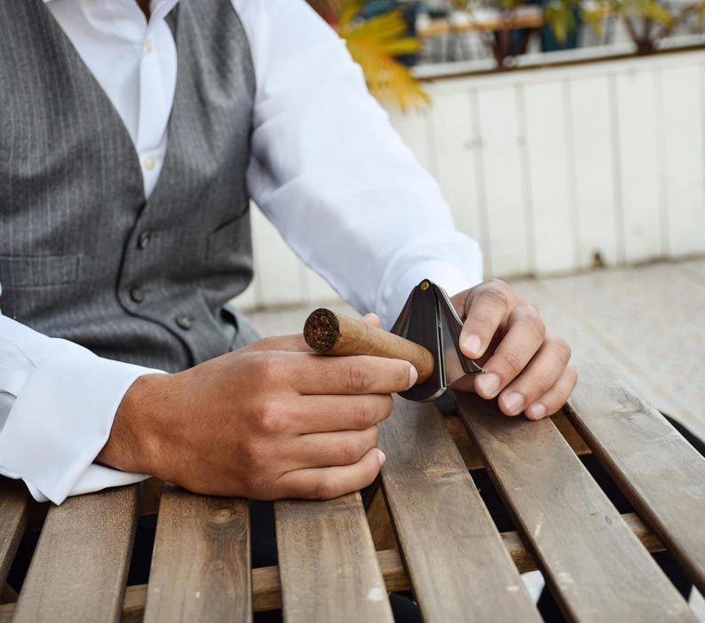 Puede incluir: Un cortapuros met&aacute;lico en forma de V con acabado plateado, colocado para cortar el extremo de un cigarro de tabaco marr&oacute;n. La herramienta presenta un dise&ntilde;o articulado con un punto de pivote central para preparar el cigarro.
