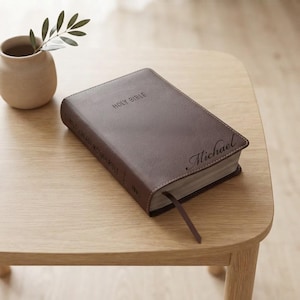 May include: A brown leather-bound Holy Bible with the name Michael engraved in script on the front cover. The book features a dark brown ribbon bookmark and the text Holy Bible embossed on the top center, with the spine labeled Foundation Study Bible.