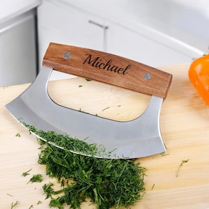 May include: A personalized wooden handled chopping knife with a stainless steel blade, resting on a wooden cutting board with chopped green herbs. The handle is engraved with the name "Michael".