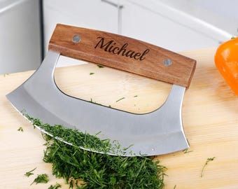 May include: A personalized wooden handled chopping knife with a stainless steel blade, resting on a wooden cutting board with chopped green herbs. The handle is engraved with the name "Michael".