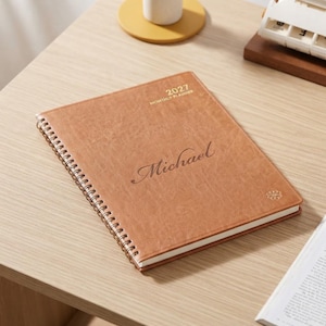 May include: A tan, spiral-bound monthly planner for the year 2027 with a textured cover. The front cover features the text 2027 MONTHLY PLANNER and the name Michael in a cursive script, along with a small circular emblem in the bottom corner.