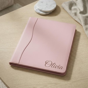 May include: A light pink rectangular portfolio folder with a smooth finish and a vertical stitched seam along the left side. The name Olivia is engraved in a dark cursive script in the bottom right corner of the front cover.