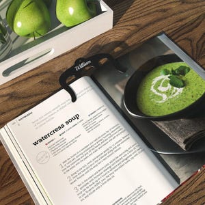May include: A close-up of a cookbook page with a recipe for watercress soup. The recipe includes instructions for preparing the soup, as well as tips for serving it. The page is open to a section titled "Watercress Soup" and features a photo of a bowl of green soup with a swirl of cream on top.