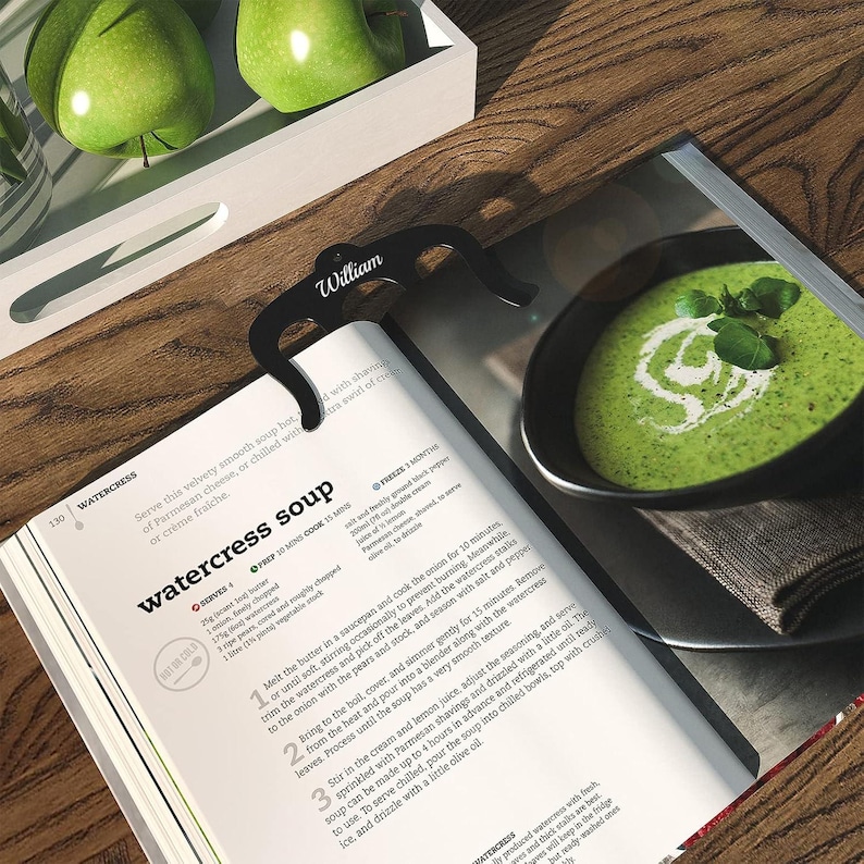 May include: A close-up of a cookbook page with a recipe for watercress soup. The recipe includes instructions for preparing the soup, as well as tips for serving it. The page is open to a section titled "Watercress Soup" and features a photo of a bowl of green soup with a swirl of cream on top.