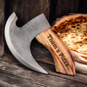 Może przedstawiać: Cutter do pizzy w kształcie siekiery z zakrzywionym ostrzem i drewnianym uchwytem. Uchwyt jest grawerowany słowami "Thom's Pizza".