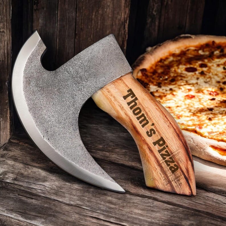 Może przedstawiać: Cutter do pizzy w kształcie siekiery z zakrzywionym ostrzem i drewnianym uchwytem. Uchwyt jest grawerowany słowami "Thom's Pizza".