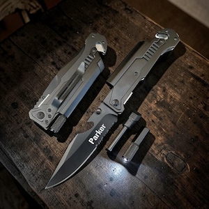 May include: A folding pocket knife with a grey metal handle and a partially serrated blade featuring the name Parker printed in white. Beside it lies a closed folding knife, a metal fire starter rod, and a small cylindrical metal flashlight.