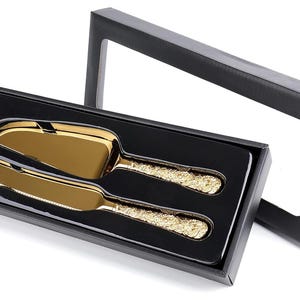 Puede incluir: Un juego de servidor de pastel y cuchillo de color dorado con mangos ornamentados, presentado en una caja de regalo negra. El juego incluye un servidor de pastel y un cuchillo, ambos con un acabado dorado brillante. La caja es negra con una ventana transparente.