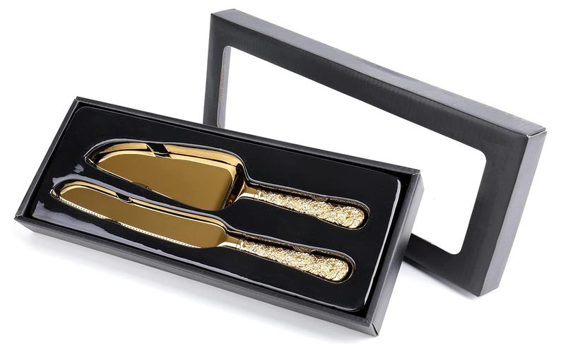 Puede incluir: Un juego de servidor de pastel y cuchillo de color dorado con mangos ornamentados, presentado en una caja de regalo negra. El juego incluye un servidor de pastel y un cuchillo, ambos con un acabado dorado brillante. La caja es negra con una ventana transparente.