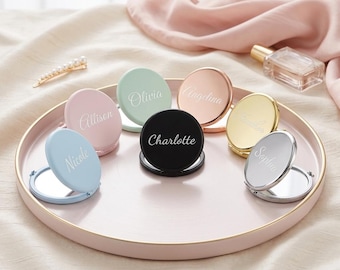 May include: A collection of seven round, foldable compact mirrors in various colours including light blue, pink, mint green, black, rose gold, gold, and silver. Each mirror features a custom name engraved in white script font on the lid.