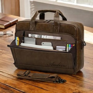 May include: A brown leather briefcase with multiple compartments, including a front flap pocket. The briefcase has a top handle, a detachable shoulder strap, and the name "Andrew" embossed on the front. It contains a newspaper, pens, and a smartphone.