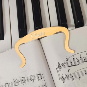 May include: Gold-colored metal page holder for sheet music with the name "Charlie" engraved on it. The page holder is on top of a sheet music book with musical notes.
