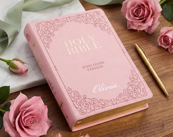 May include: A pink Holy Bible with gold gilded edges, the cover features the text "Holy Bible" and "King James Version". The name "Olivia" is printed on the cover. The Bible is surrounded by pink roses and a gold pen on a wooden surface.