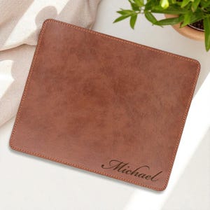 May include: A rectangular brown leather mousepad with stitched edges. The name "Michael" is elegantly engraved in a cursive font on the lower right corner. The mousepad is a solid color and has a smooth texture.