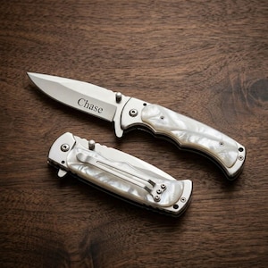 May include: A folding pocket knife with a silver blade engraved with the name Chase and a handle featuring a pearlescent white finish. A second closed folding knife with a matching pearlescent handle and a silver metal pocket clip is positioned below.
