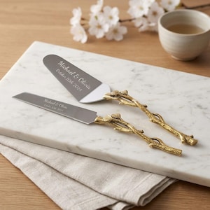 May include: A silver-toned cake knife and server set with gold-colored metal handles shaped like textured tree branches. The blades are engraved with the text Michael & Olivia and October 10th 2024. The set features a serrated edge on the knife.