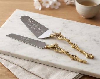 May include: A silver-toned cake knife and server set with gold-colored metal handles shaped like textured tree branches. The blades are engraved with the text Michael & Olivia and October 10th 2024. The set features a serrated edge on the knife.