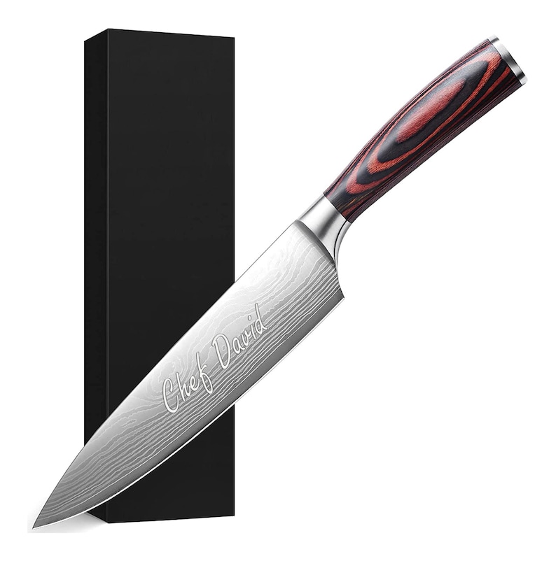 Puede incluir: Un cuchillo de chef de acero inoxidable con una hoja estampada y un mango de madera con tonos rojos y marrones. El texto "Chef David" est&aacute; grabado en la hoja. Una caja negra est&aacute; en el fondo.