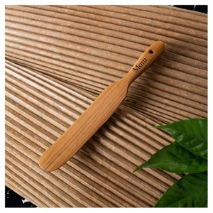 May include: A wooden spatula with a light brown finish, engraved with the word "Mom" on the handle. The spatula has a flat, rectangular blade and a small hole at the end of the handle. The spatula is lying on a textured, brown surface.