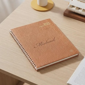 May include: A tan, spiral-bound monthly planner with a textured cover. The front cover features the text 2027 MONTHLY PLANNER at the top and the name Michael in a cursive script font in the centre, with a small decorative floral icon.