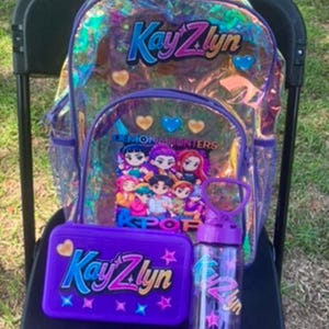 Könnte beinhalten: Ein durchsichtiger, irisierender Rucksack mit violetten Rändern, der den Namen KayZlyn und eine Grafik von Anime-Figuren mit dem Text Demon Hunters KPOP zeigt. Enthält ein passendes violettes Federmäppchen und eine violette Trinkflasche, beide mit KayZlyn beschriftet.