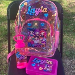 May include: A clear iridescent backpack with pink trim, a matching pink water bottle, and a pink lunch box. Each item is personalized with the name Layla in blue and purple gradient text. The backpack and lunch box feature K-pop themed graphics.
