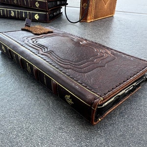May include: A brown leather phone case designed to look like a vintage book. The case features embossed detailing, gold accents, and a small leather tab closure. Additional cases are visible in the background.