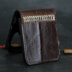 May include: A brown leather wallet with a stitched design and a black lanyard. The wallet is made of textured leather and has a cream-coloured stitch detail across the top. The wallet is standing upright.