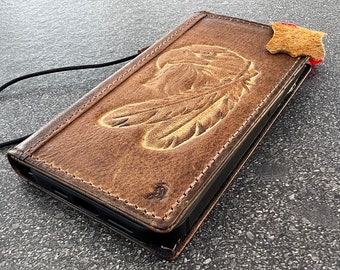 May include: A brown leather phone case with a detailed embossed design of a Native American headdress with feathers. The case has a black strap and a brown leather flap.