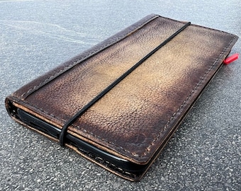 May include: A brown leather phone case with a black elastic band closure. The case has a distressed, vintage look.
