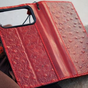 May include: Red leather phone case with a textured finish and red stitching. The case features a black frame for the phone and a folding flap for screen protection. The case is open, showing the phone's camera.