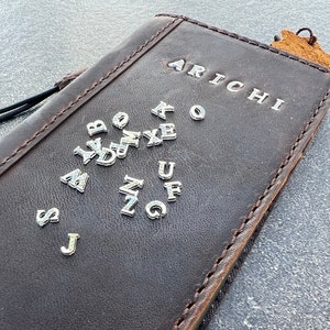 Könnte beinhalten: Ein braunes Lederjournal mit dem Wort "ARICHI" geprägt auf der Vorderseite. Das Journal hat eine Vielzahl von silbernen Metallbuchstaben, die an der Vorderseite angebracht sind, darunter die Buchstaben A bis Z.
