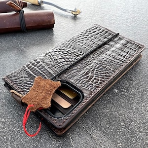 May include: A brown leather phone case with a crocodile pattern. The case has a flap that folds over the phone and a slot for cards. The case is lying on a gray surface.