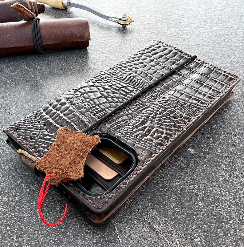 May include: A brown leather phone case with a crocodile pattern. The case has a flap that folds over the phone and a slot for cards. The case is lying on a gray surface.