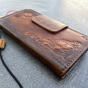 May include: A brown leather phone case with a floral design and a flap closure. The case has a strap attached to it.