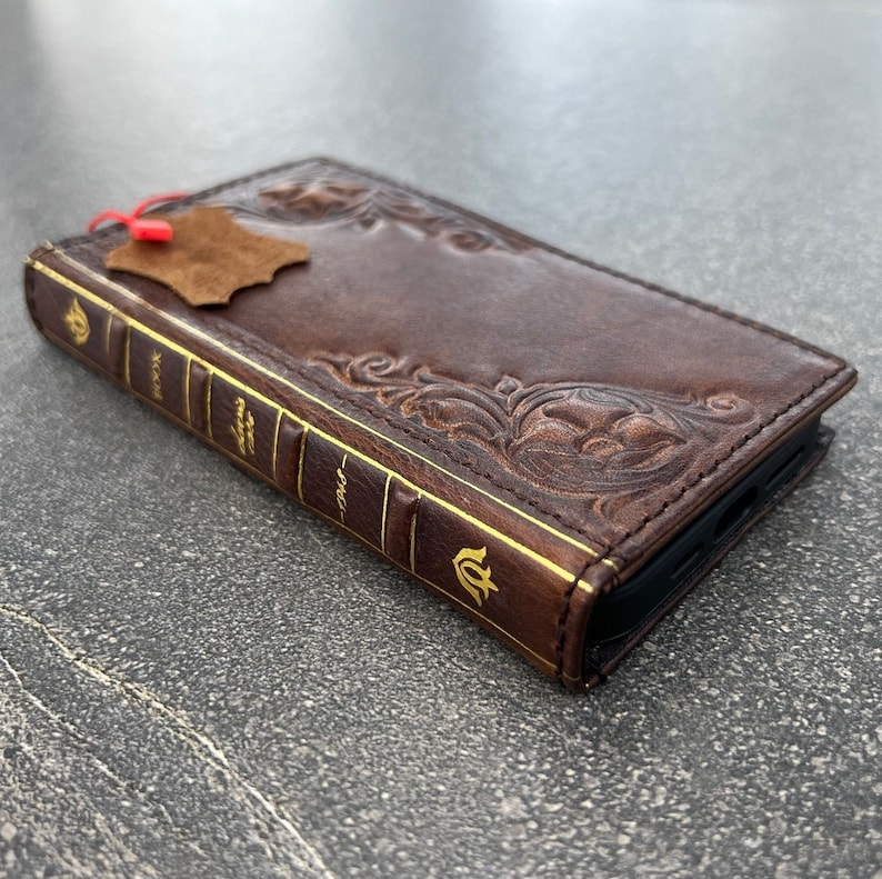 May include: A brown leather phone case designed to look like a vintage book. The case features embossed floral designs and gold lettering on the spine that reads "BOOK 1946". A brown leather tag is attached with a red string.