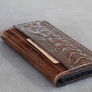Könnte beinhalten: Eine braune Leder-Handyhülle mit einer strukturierten Straußenlederklappe. Die Hülle hat weiße Nähte und einen kleinen Kartenhalter aus Holz. Ein Lederriemen ist an der Seite befestigt.
