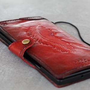 Könnte beinhalten: Rote Leder-Handyhülle mit geprägtem Federdesign. Sie hat einen Druckknopfverschluss mit goldenem Knopf und einen schwarzen Riemen. Die Hülle ist für ein Mobiltelefon konzipiert.