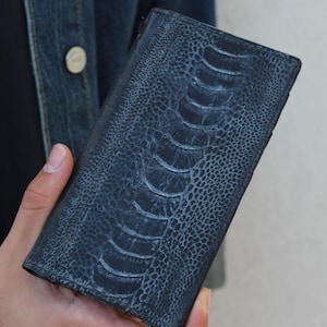 Puede incluir: Cartera de cuero azul oscuro con un patrón de pata de avestruz. La cartera rectangular tiene un cierre de cremallera. La cartera está en la mano de una persona.