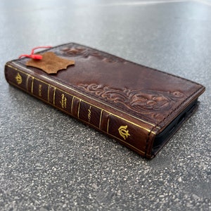 May include: A brown leather phone case designed to look like a vintage book. The case features embossed floral patterns and gold lettering on the spine, with the words "BOOK" and "CLASSIC CASE" visible. A small leather tag hangs from the top.
