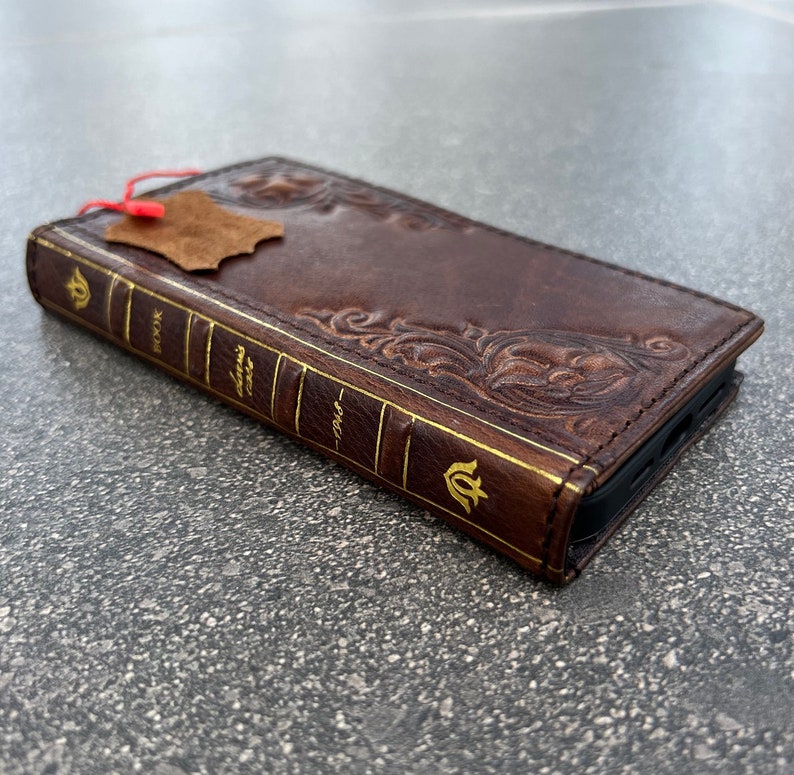 May include: A brown leather phone case designed to look like a vintage book. The case features embossed floral patterns and gold lettering on the spine, with the words "BOOK" and "CLASSIC CASE" visible. A small leather tag hangs from the top.