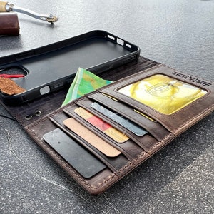 May include: A brown leather wallet case for a smartphone with a black phone case inside. The wallet has multiple card slots and a flap with a clear window for displaying an ID card. The wallet is open and the phone case is visible.