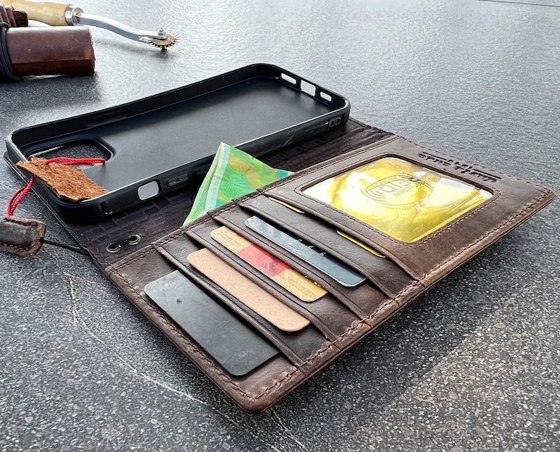 May include: A brown leather wallet case for a smartphone with a black phone case inside. The wallet has multiple card slots and a flap with a clear window for displaying an ID card. The wallet is open and the phone case is visible.