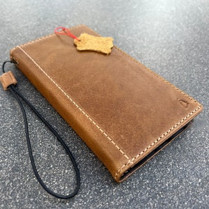 May include: A brown leather phone case with a strap. The case has a stitched edge and a small leather tag attached.