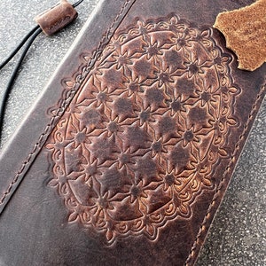 May include: Dark brown leather wallet with an embossed floral pattern. The wallet has a stitched border and a black cord with a leather loop. A small piece of brown leather is attached to the wallet.