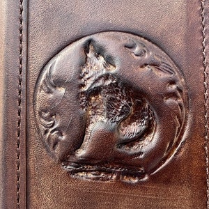 Op de afbeelding: Close-up van een bruin lederen item met een cirkelvormig reliëfontwerp. Het ontwerp toont een gedetailleerde afbeelding van een wolf die naar de maan huilt, omringd door decoratieve elementen. Het leer heeft een getextureerd uiterlijk.