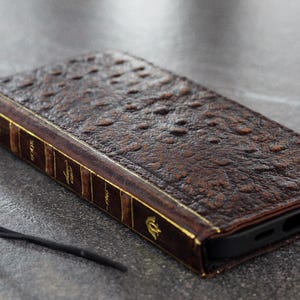 Puede incluir: Funda de teléfono de cuero marrón con diseño de libro antiguo. La funda tiene una superficie texturizada, detalles dorados y un cordón negro. La funda está diseñada para guardar un smartphone.